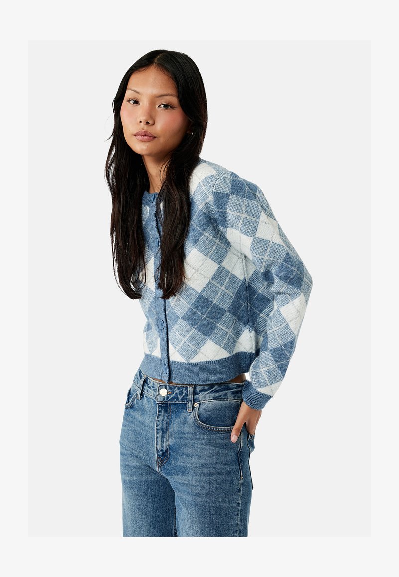 Joven mujer que lleva un cárdigan abotonado de rombos azul y blanco y vaqueros azules, posando con una mano en el bolsillo frente a un fondo liso.