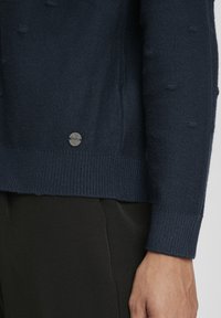 Pull bleu marine avec un tricot texturé, ourlet et poignets côtelés, et un accent de logo métallique près du bas, associé à un pantalon noir texturé.