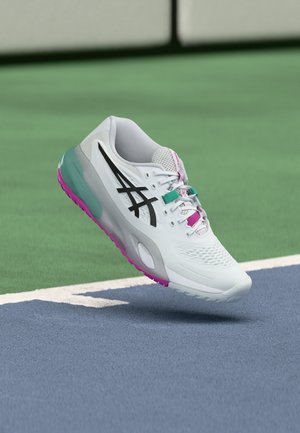 Zapatilla deportiva blanca con logo negro y detalles en rosa y verde azulado, suspendida sobre la superficie azul y verde de una pista de tenis.