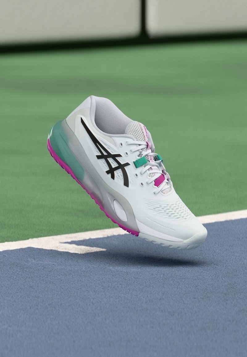 Λευκό αθλητικό παπούτσι με μαύρο λογότυπο, ροζ και τιρκουάζ λεπτομέρειες, να αιωρείται πάνω από μια επιφάνεια γηπέδου τένις σε μπλε και πράσινο χρώμα.