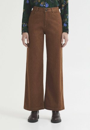 Femme portant un pantalon marron taille haute à jambes larges, des bottes noires et un chemisier vert avec un imprimé floral bleu et jaune, debout sur un fond blanc.