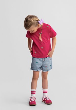Młoda dziewczyna z warkoczami związanymi fioletową kokardką, ubrana w czerwoną koszulkę w wzór z wisienkami, dżinsowe szorty z haftem serc oraz różowe trampki.