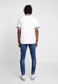 White short-sleeve shirt with black stripe accents, paired with blue denim jeans, standing against a neutral background.