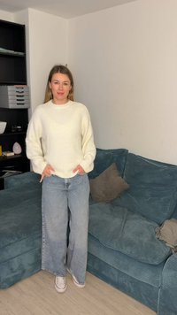 Femme debout à l'intérieur portant un pull blanc, un jean large bleu clair et des baskets blanches à côté d'un canapé d'angle bleu sarcelle avec des coussins.