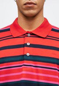 Άνδρας με ορατό λαιμό και κάτω μέρος προσώπου που φοράει κόκκινη πόλο μπλούζα με οριζόντιες ρίγες σε ναυτικό, ροζ και πράσινο χρώμα και τρία κουμπιά κουμπωμένα.