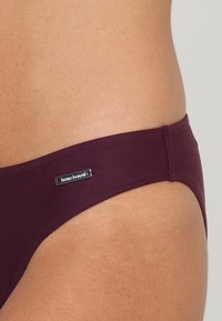 Burgunderfarbene Bikini-Hosen aus dehnbarem Stoff mit glatter Textur, ausgestattet mit einem kleinen metallischen Markenschild an der Seite und einem minimalen Bedeckungsdesign.