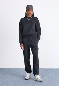 Sort sweatshirt og sweatpants i blødt stof, med et broderet logo. Sammen med en grå kasket og grå løbesko med hvide detaljer.