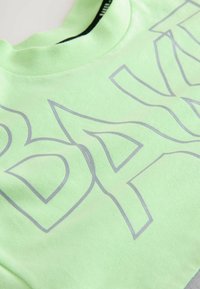 Sudadera verde claro con cuello redondo, que presenta un gran texto plateado en negrita. La tela tiene una textura suave y un ajuste relajado.