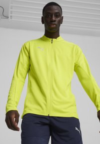 Puma Zip-Jacke in leuchtendem Gelb, aus glattem Material, mit langen Ärmeln und kleinem Logo auf der Brust.