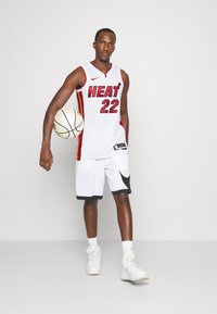 Baltā basketbola forma ar sarkanām un dzeltenām akcentiem, uz kuras rakstīts 'HEAT' un '22'. Kombinēta ar melni izstrādātām baltām šortiem un baltām sporta apaviem.
