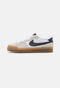 Beige ruskind og hvid mesh sneakers med sorte accenter, snørrebånd design og en gennemsigtig brunt gummisål. Nike-logo på siden.
