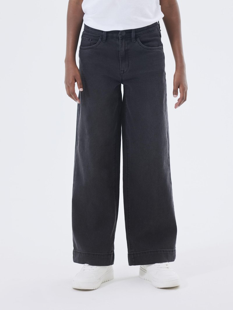 Jeans de denim negro de pierna ancha con una cintura alta, cinco bolsillos y dobladillos enrollados, combinados con zapatillas blancas sobre un fondo neutro.