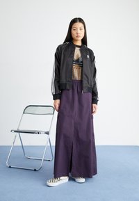 Black satin bomber jacket with white stripes, mesh top, purple midi skirt with side slits, and checkered slip-on shoes. Clear chair in background.