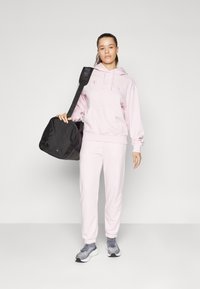 Lys rosa hoodie med snore, matchende sweatpants og grå sportssko. Sort duffelbag med skulderrem i hånden. Enkel design.