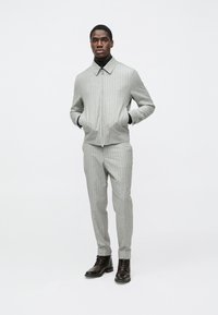 Svetlo siva jakna z enakomernimi črtami, ovratnikom in zadrgo, združena z ujemajočimi hlačami in črnimi škornji z vezalkami, s sijočo teksturo.