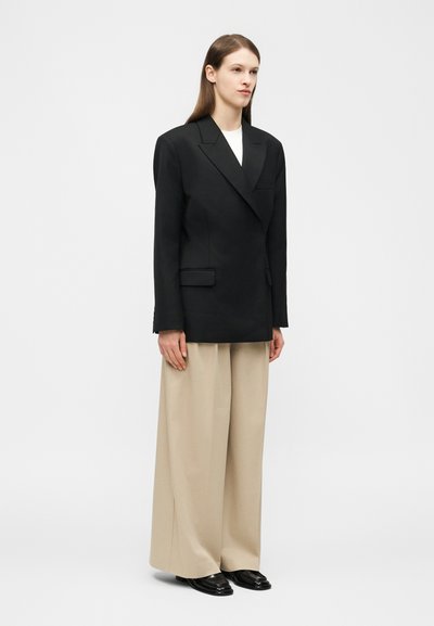 Osoba v nadrozmernom čiernom sakom, bielom topuse, širokých béžových nohaviciach a čiernych topánkach stojaca pred jednoduchým svetlým pozadím.