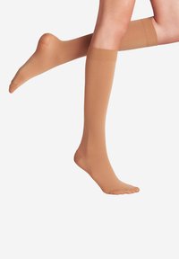 Gambe di donna che indossano calze a compressione beige fino al ginocchio, una gamba piegata all'indietro all'altezza del ginocchio su sfondo bianco.