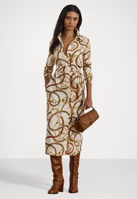 Vestito lungo con bottoni, caratterizzato da un motivo a catena in tonalità crema, rosso e oro. Presenta una vita rimovibile, tasche ed è abbinato a una borsa marrone e stivali.
