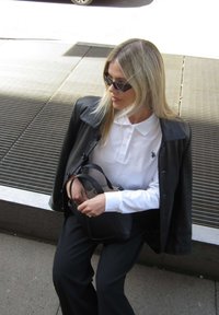 Blondīne ar melnām saulesbrillēm, melnu ādas jaku pār balu krekla apkakli, turot melnu somiņu, sēžot uz āra metāla režģa.