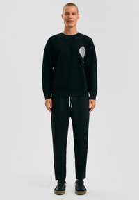 Zwarte gebreide trui met een gestructureerd schelpdesign op de borst, gecombineerd met zwarte jogginbroeken en zwarte sportschoenen.