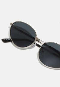 Ochelari de soare rotunzi cu lentile închise, cadru din metal argintiu, ușor, și brațe din plastic negru. Prezintă pernuțe ajustabile pentru nas.