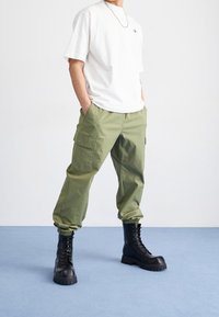Person som bär en vit oversized t-shirt, olivgröna cargo-byxor med elastiska manschetter och svarta kängor med snörning, stående på en blå golv.