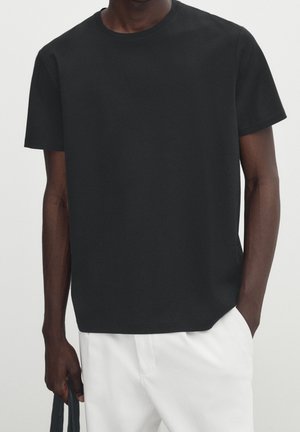 Homme portant un t-shirt noir uni à manches courtes rentré dans un pantalon blanc, une main dans la poche et tenant un sac noir de l'autre.