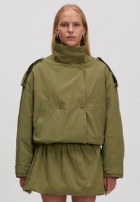 Olijfgroene jas met een hoge kraag en dubbelknopen. Heeft volumineuze mouwen en een gather taille. Gelaagd materiaal.