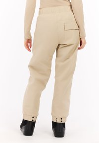 Beige, hoge taille broek met elastische tailleband en details van zijzakken. Omgeslagen zomen met drukknopen en zwarte schoenen. Gladde textuur.