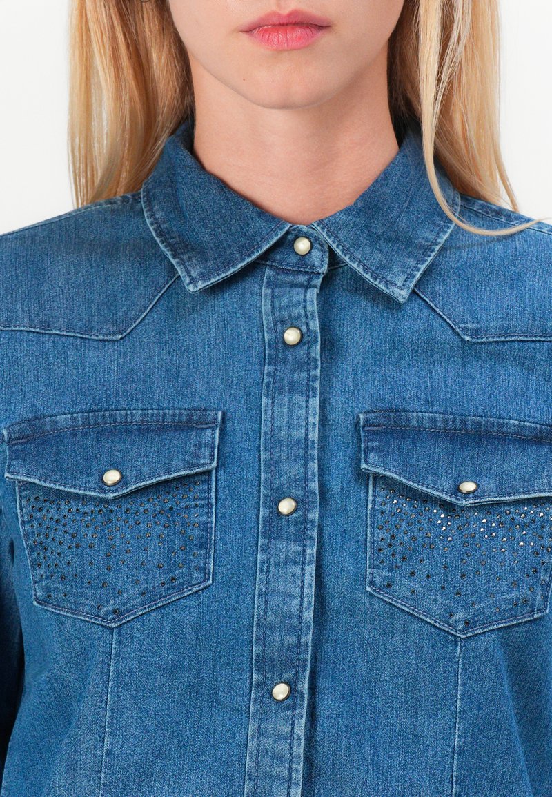 Camisa de denim en azul medio con cuello clásico, dos bolsillos delanteros y/details decorativos de tachuelas en los bolsillos. Acabado con botones a presión metálicos.