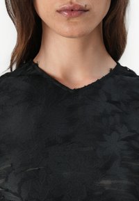 Svart teksturert topp med blomstermønster, med V-utringning og frynsete kanter. Stoffet ser lett og semi-transparent ut.