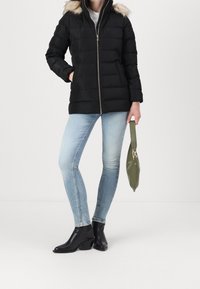 Zwart gewatteerd jack met een capuchon van nepbont en een ritssluiting aan de voorkant, gecombineerd met lichtblauwe skinny jeans en zwarte enkellaarsjes. Draagt een groene handtas.