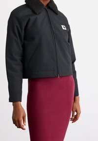 Zwarte cropped jas met een getextureerde zwarte shearlingkraag, rits aan de voorkant en zijzakken. Gecombineerd met een geribde bordeauxrode rok.