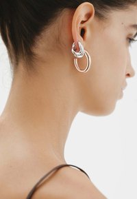 Pendientes de aro de metal plateado y rosa que presentan lazos entrelazados, exhibidos en una oreja sobre un fondo neutro.