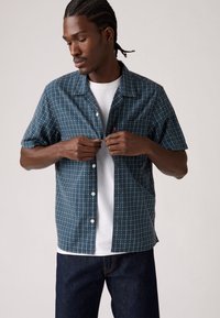 Korte mouw shirt in donker teal stof met een ruitpatroon in wit en rood. Met knoopsluiting ontwerp, gedragen over een wit T-shirt met ronde hals.