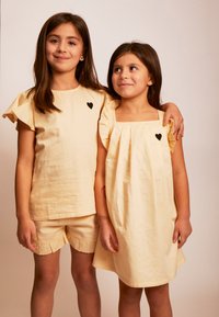 Zwei Mädchen tragen hellgelbe Kleidung. Eines trägt ein Rüschentop mit einem schwarzen Herz und passende Shorts. Das andere trägt ein ärmelloses Kleid mit einem Herz.