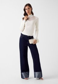 Witte trui met doorzichtig borstpaneel, gecombineerd met marineblauwe jeans met brede pijpen en contrasterende denimmanchetten. Model houdt een crème clutch vast.