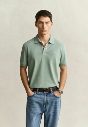 Junger Mann mit kurzen dunklen Haaren, der ein hellgrünes Polohemd, blaue Jeans, braunen Gürtel und eine Armbanduhr trägt, steht mit den Händen in den Taschen.