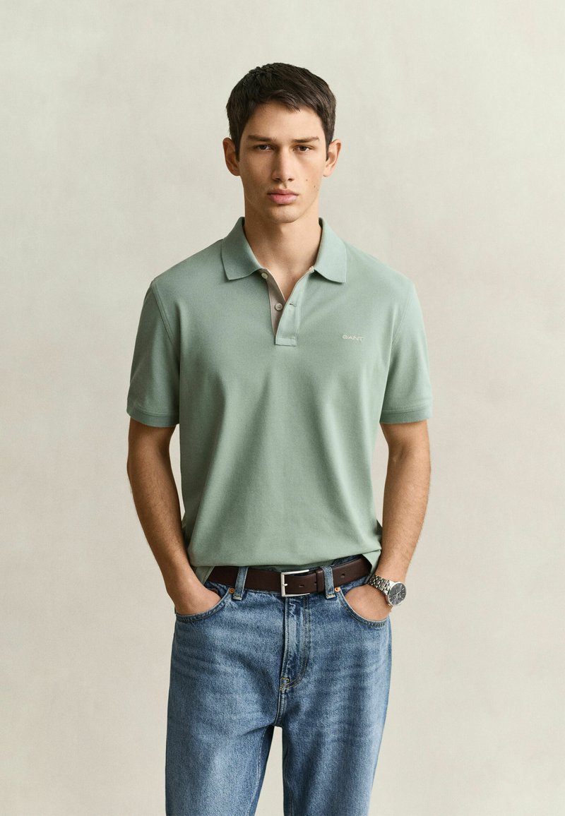 Jeune homme aux cheveux courts et foncés portant un polo vert clair, un jean bleu, une ceinture marron et une montre-bracelet, debout les mains dans les poches.