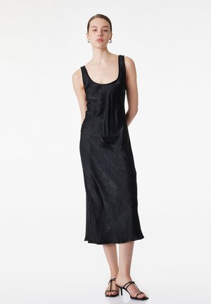 Joven mujer con vestido midi negro sin mangas y textura sutil, usando sandalias negras de tacón con tiras, de pie con las manos detrás de la espalda.