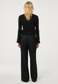 Zwarte gebreide trui met lange mouwen gecombineerd met zwarte broek met hoge taille, voorzien van witte verticale krijtstrepen. Effen achtergrond.