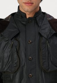 Wasserabweisende schwarze Jacke mit hohem Kragen, fünf großen Knöpfen und zwei mit Reißverschluss versehenen Taschen. Mit einem strukturierten Cord-Futter an den Schultern.