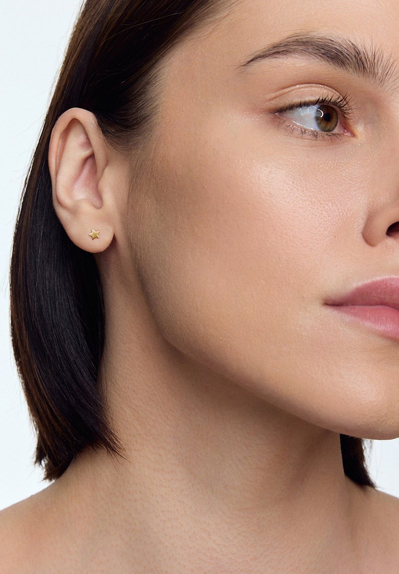 Złota gwiazda wkrętka noszona na lewym uchu. Kolczyk jest mały, ma płaską powierzchnię i miękki blask, doskonale pasując do gładkiej skóry.
