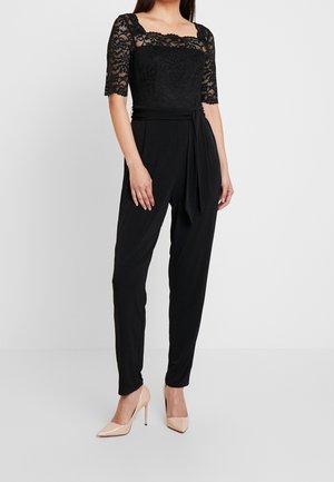 Mono de encaje negro con cuello cuadrado y mangas tres cuartos. Presenta un cuerpo ajustado, pantalones fluidos y un cinturón para atar a la cintura. Se complementa con tacones beige.