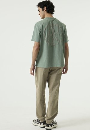 Mann steht abgewandt, trägt ein grünes T-Shirt mit großem kreisförmigen Logo auf dem Rücken, beige Hose und schwarz-weiße Sneaker.