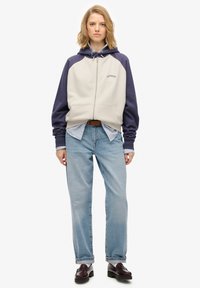 Giacca con cappuccio, con corpo beige e maniche blu scuro, dotata di tasche frontali e cerniera, indossata sopra una camicia a righe blu e bianche e jeans chiari.