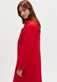 Czerwony wełniany płaszcz z wyciętym kołnierzem i długimi rękawami, o prostym kroju i bocznymi kieszeniami, prezentowany na jednolitym tle.