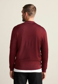 Vínový pletený sveter s dlhými rukávmi, výstrihom do V a žebrovanými manžetami. Látka pôsobí hladko a mierne priliehavo, nosená cez bielu košeľu.