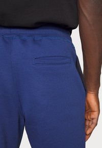 Blue sweatpants with a smooth texture, featuring a back pocket and contrasting stitching. Elastic waistband for comfort and fit.