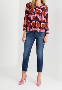 Blouse multicolore à manches longues avec des motifs circulaires abstraits en rose, orange et marron ; associée à un jean en denim délavé et des talons roses.